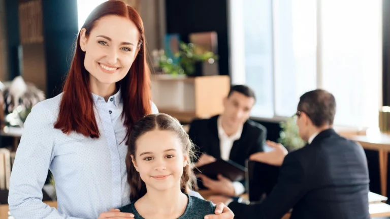 Professional woman with red hair and young girl smiling in office, two men conversing in background.