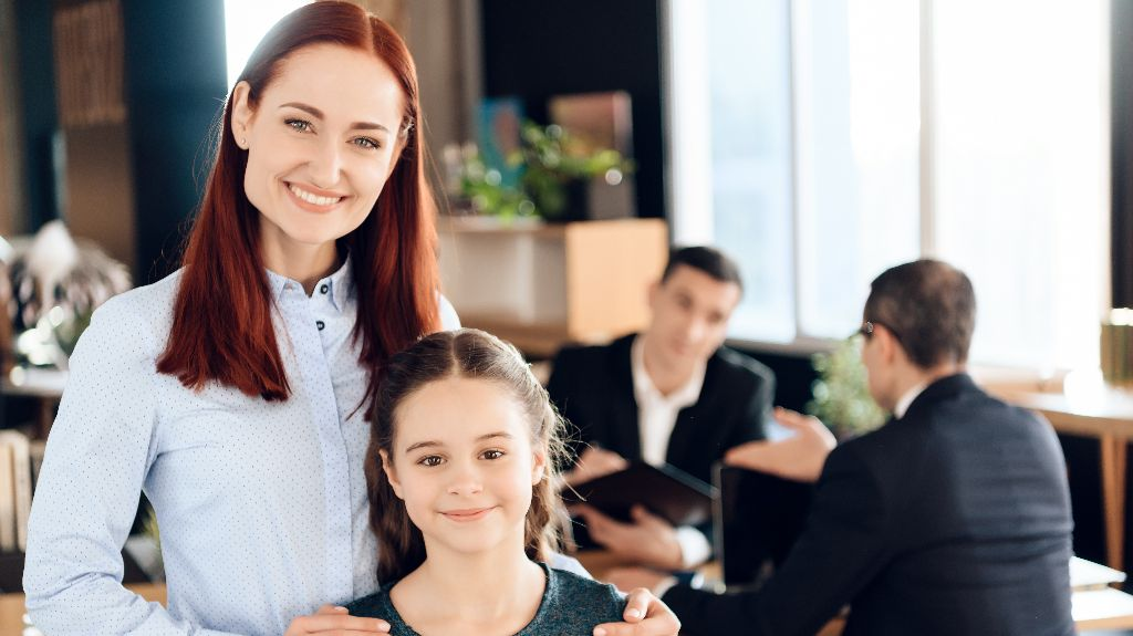 Professional woman with red hair and young girl smiling in office, two men conversing in background.
