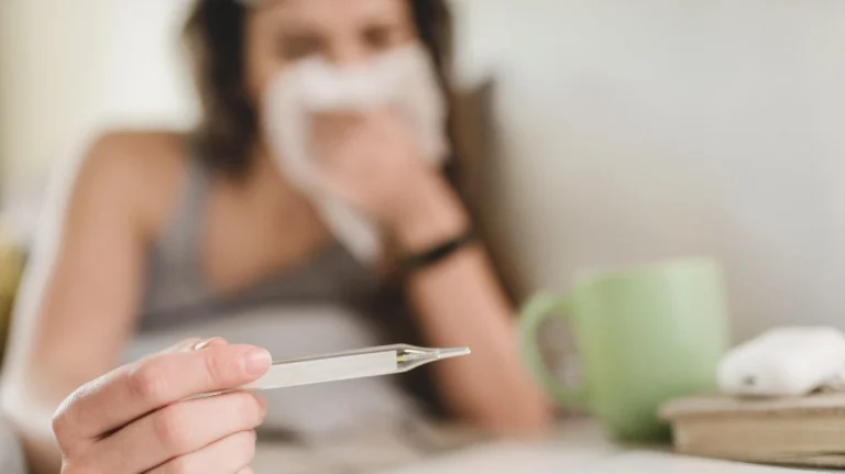 Person holding a thermometer with a tissue near their face and a green mug on the table.