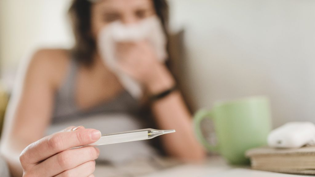Person holding a thermometer with a tissue near their face and a green mug on the table.