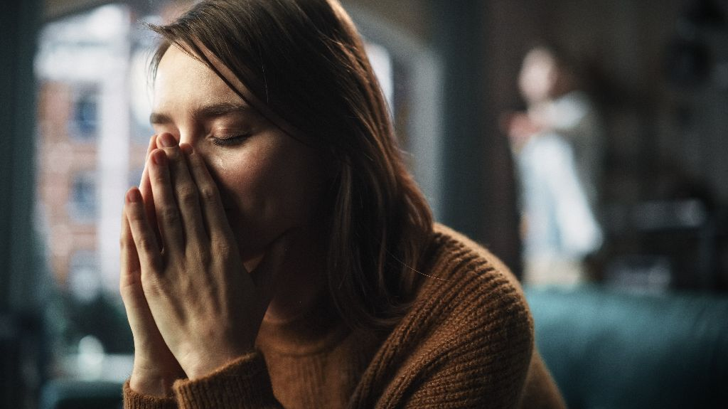 Young woman in brown sweater with eyes closed, hands covering face, indoors.