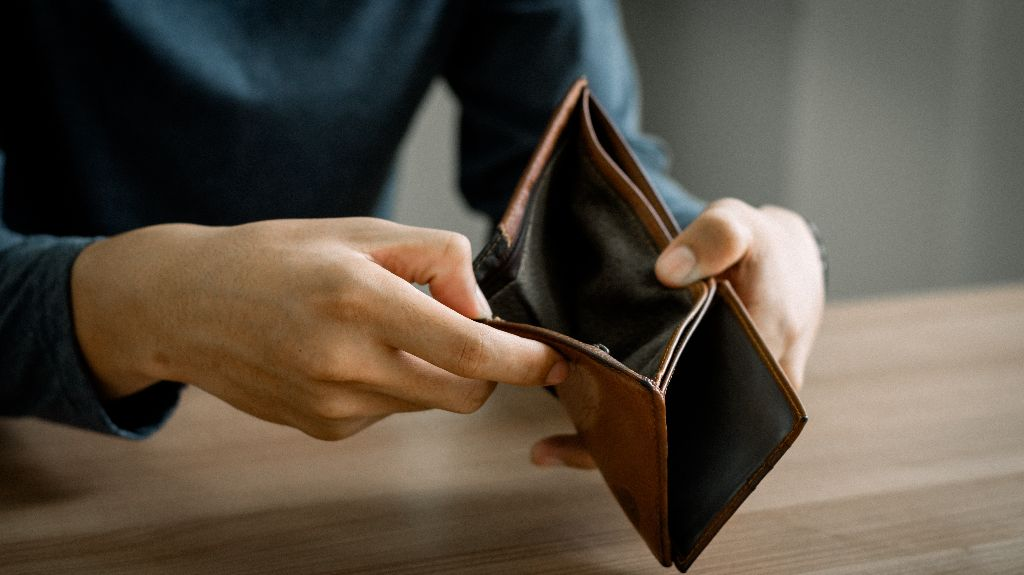 Person wearing a blue long-sleeve shirt holding an empty brown leather wallet over a wooden table.