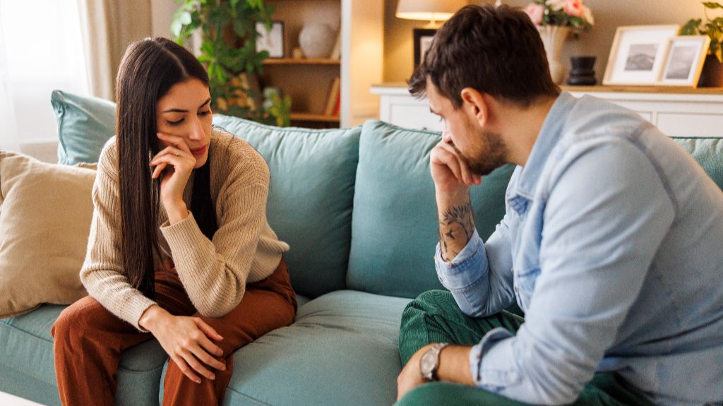 Two adults, a woman and a man, sitting on a teal couch in a living room, both appearing contemplative.