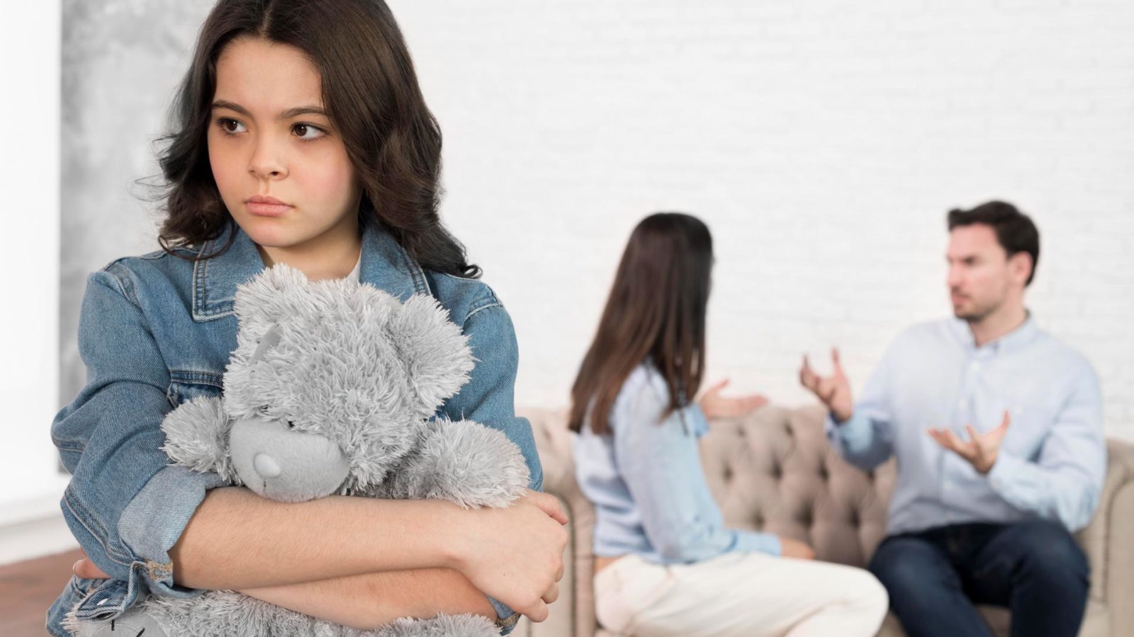 child holding a teddy bear while couple fights in background