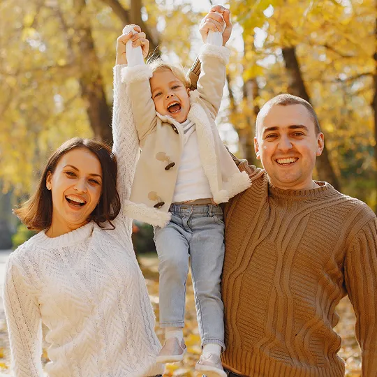 Smiling family outdoors in autumn, parents lifting young child between them.