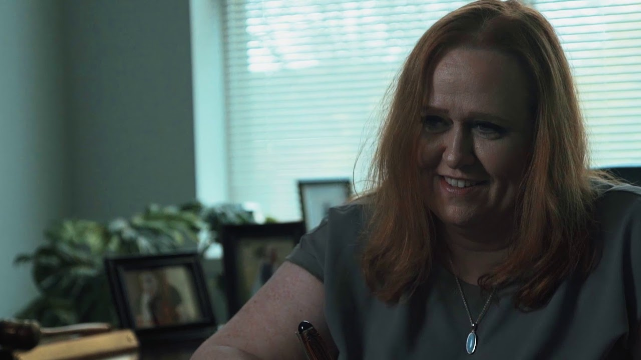 Middle-aged woman with red hair smiling in an office setting with framed photos and plants.