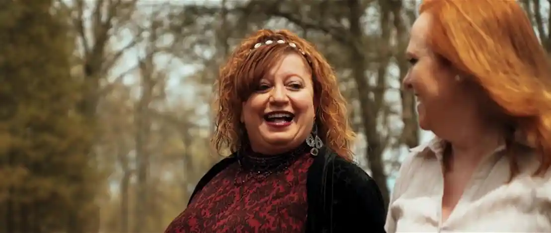 Two middle-aged women with red hair conversing outdoors in a wooded area.
