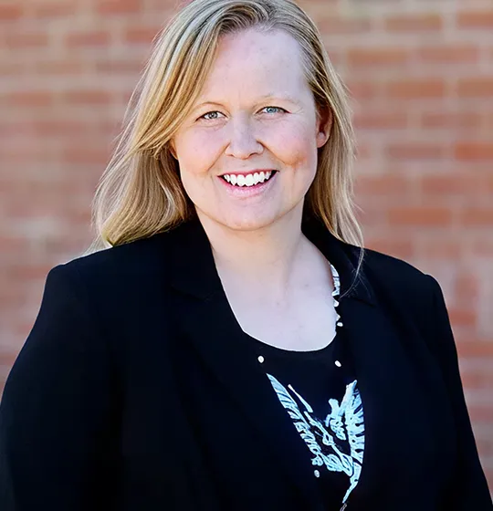 Professional portrait of a smiling woman with blonde hair, wearing a black blazer against a brick wall background.