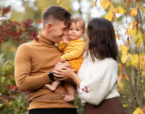 A man and woman holding a baby outdoors amid autumn foliage.