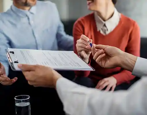 Two individuals reviewing a document on a clipboard while a third person hands over a pen.