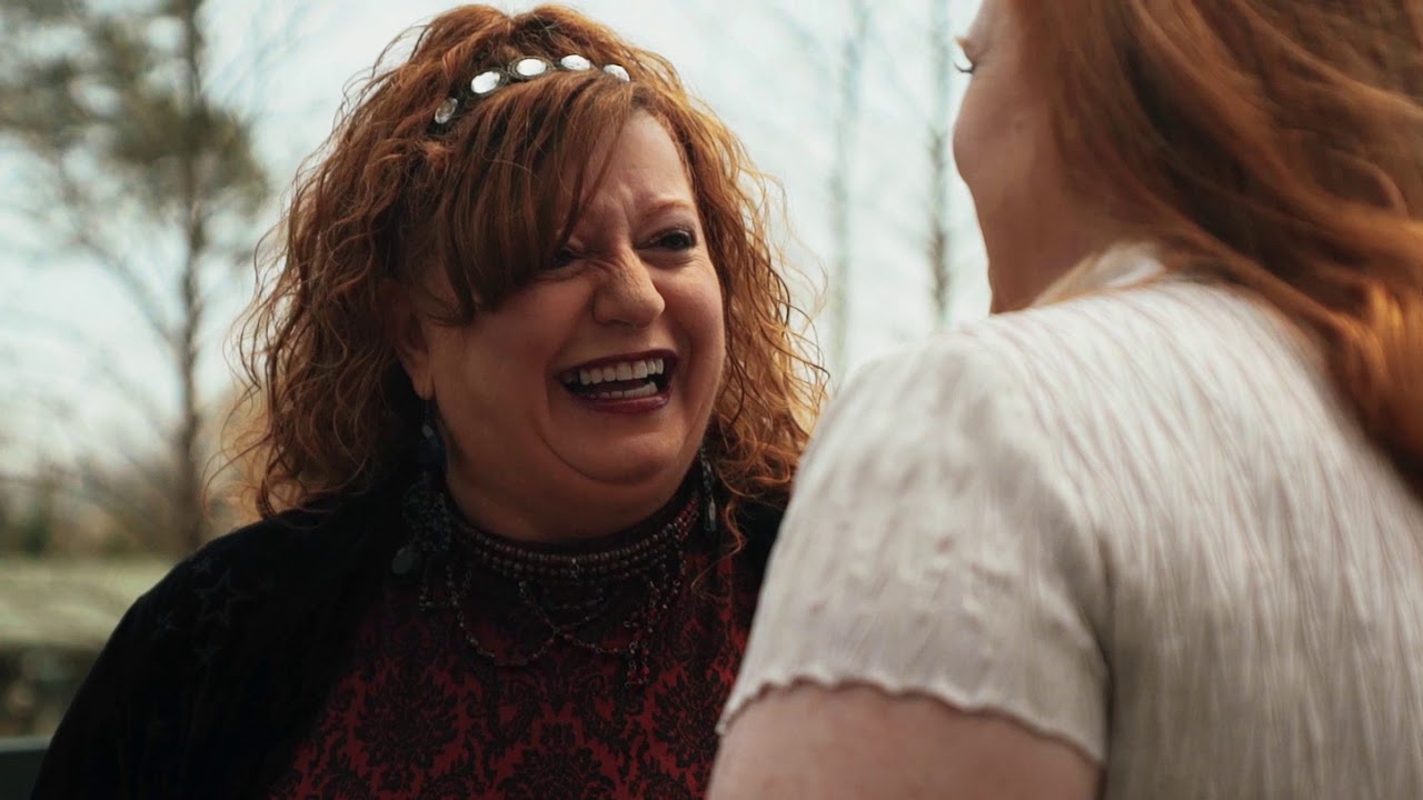 Two women engaged in conversation outdoors, one smiling broadly with curly red hair and a black and red patterned top.