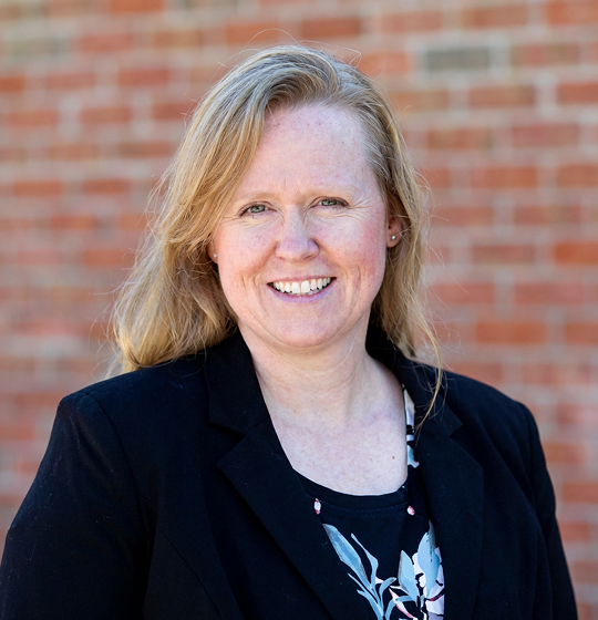 Professional portrait of a smiling woman with blonde hair, wearing a black blazer and floral top, standing against a brick wall background.