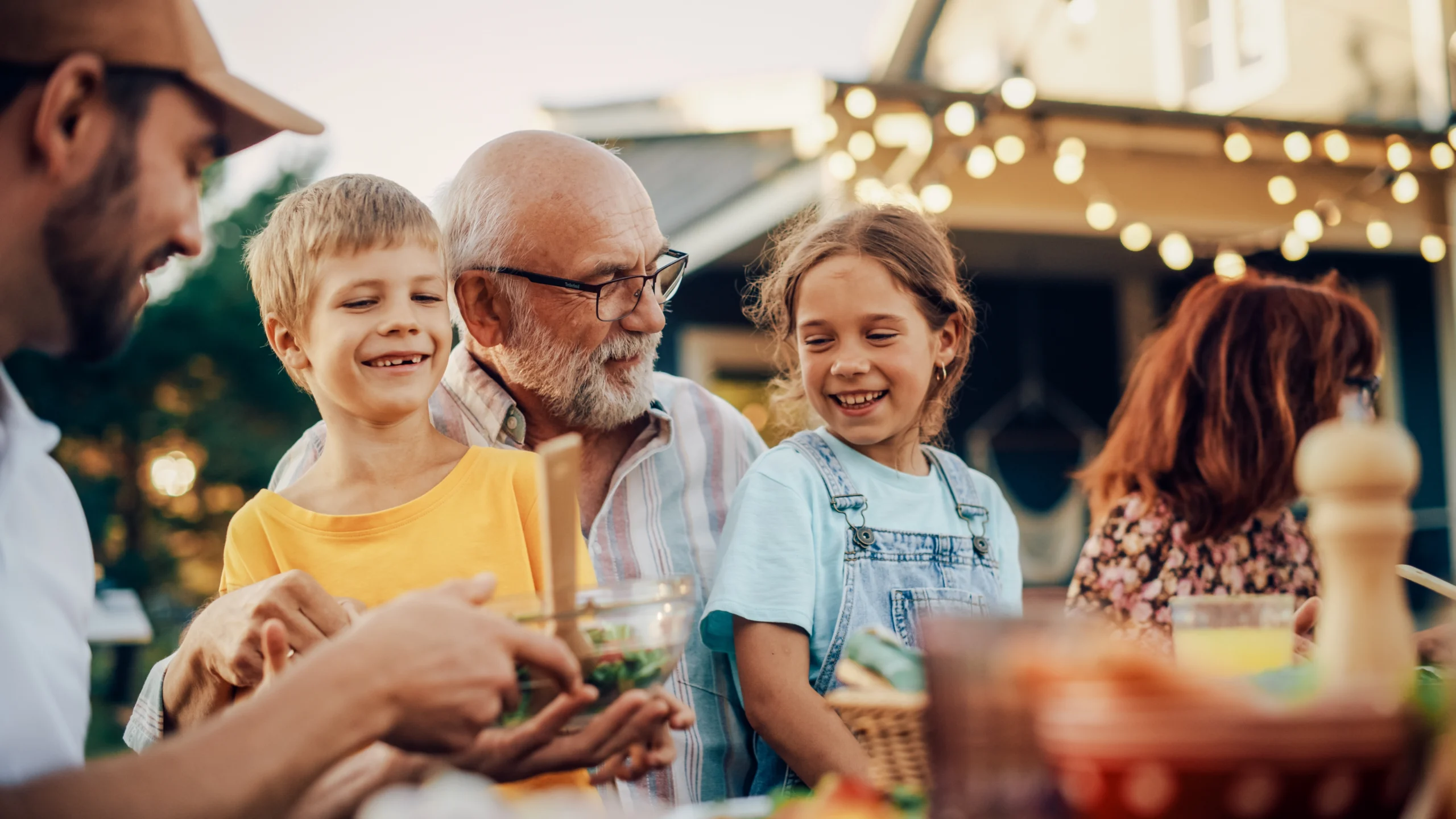 Multi-generational family enjoying outdoor meal with warm string lights in background.