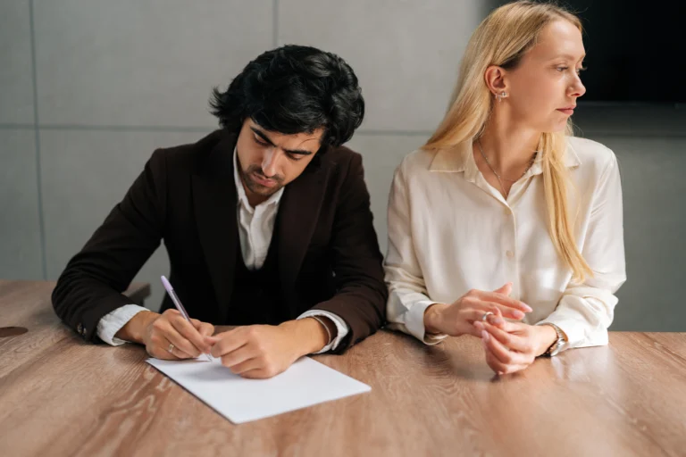 Two professionals seated at a wooden table, a man writing and a woman looking away.