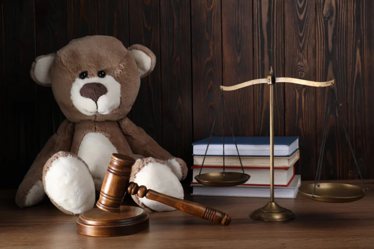 Brown teddy bear, wooden gavel, brass scales of justice, and stacked books on wood table.