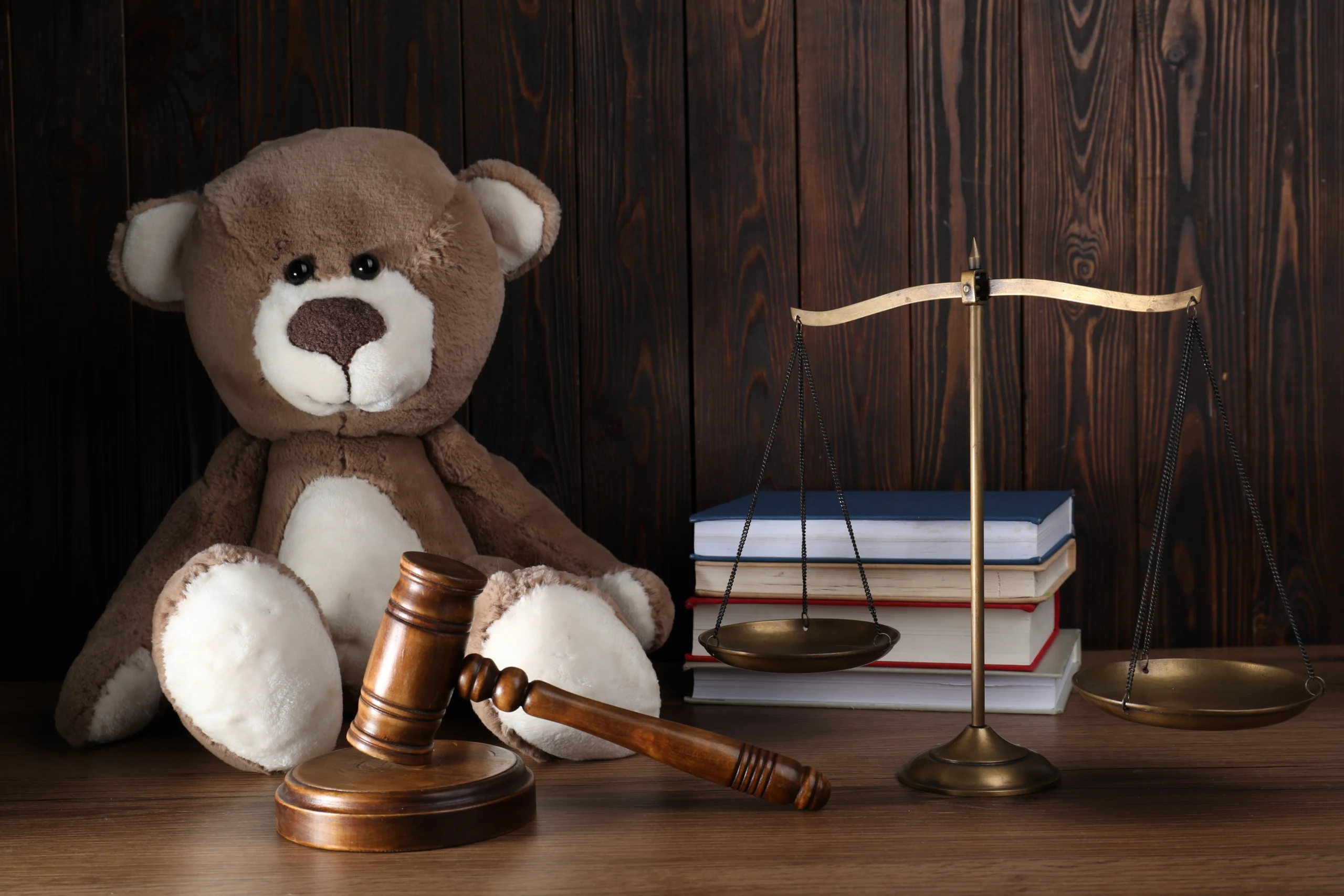 Brown teddy bear, wooden gavel, brass scales of justice, and stacked books on wood table.