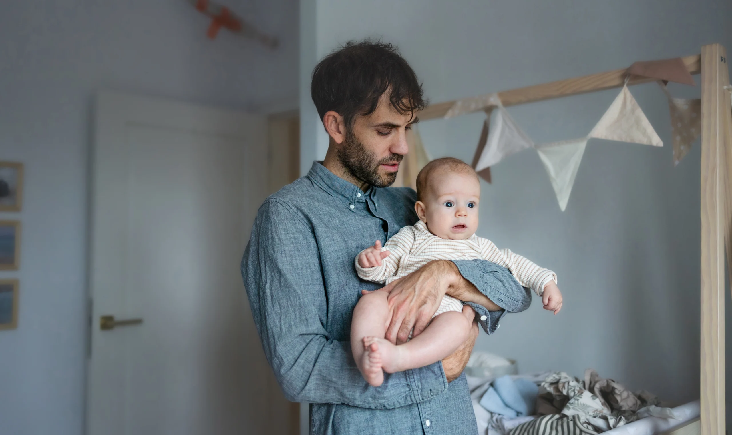 Man in a gray shirt holding a baby in a nursery with soft-colored decorations.