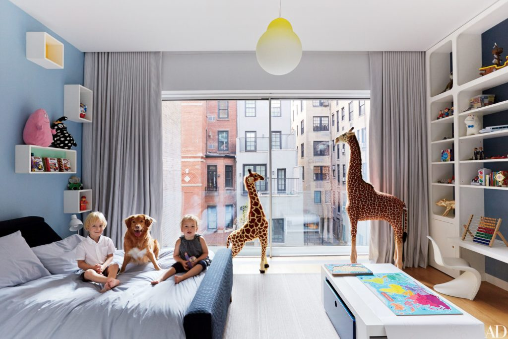 Children and dog sitting on bed in modern bedroom with large window and giraffe toys.
