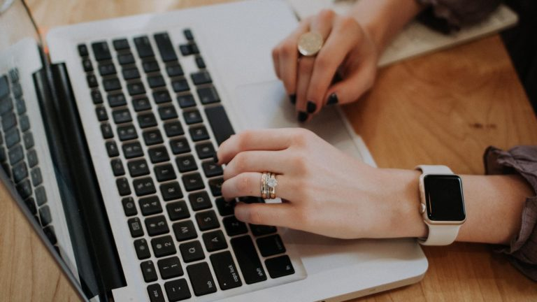 Close-up of hands typing on a laptop keyboard, wearing rings and a smartwatch.