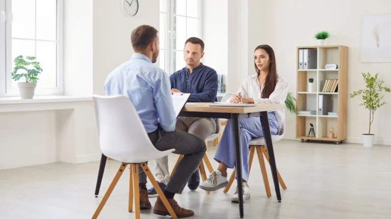Three professionals engaged in a discussion around a wooden table in a bright office.