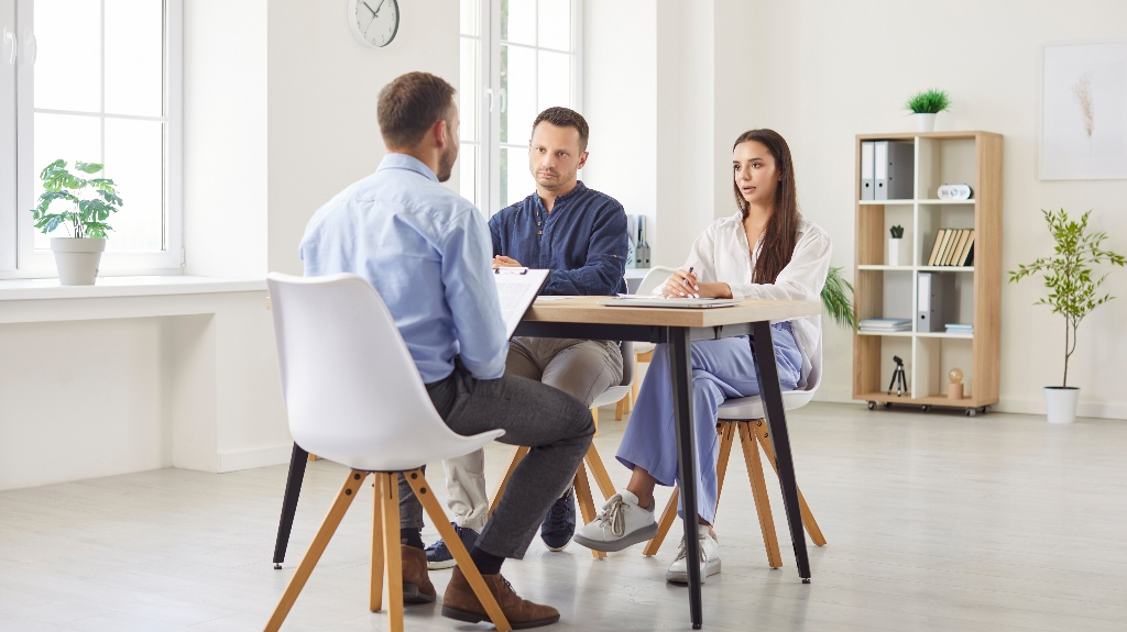 Three professionals engaged in a discussion around a wooden table in a bright office.