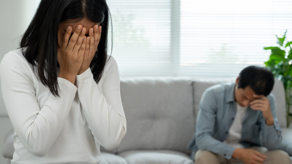 Woman covering her face with hands in foreground, man sitting on couch holding head in background.