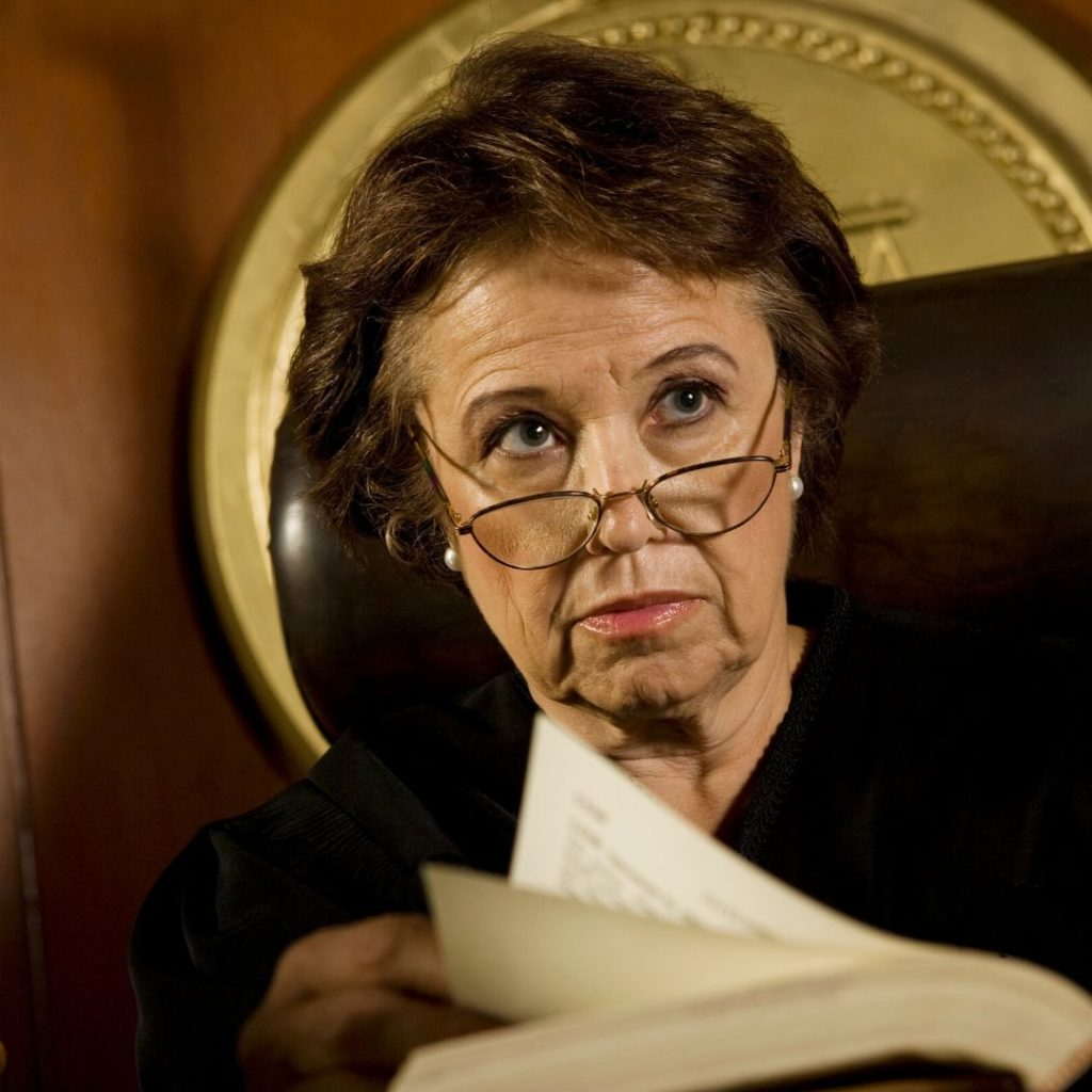 Middle-aged female judge with glasses reading a document, seated in courtroom.