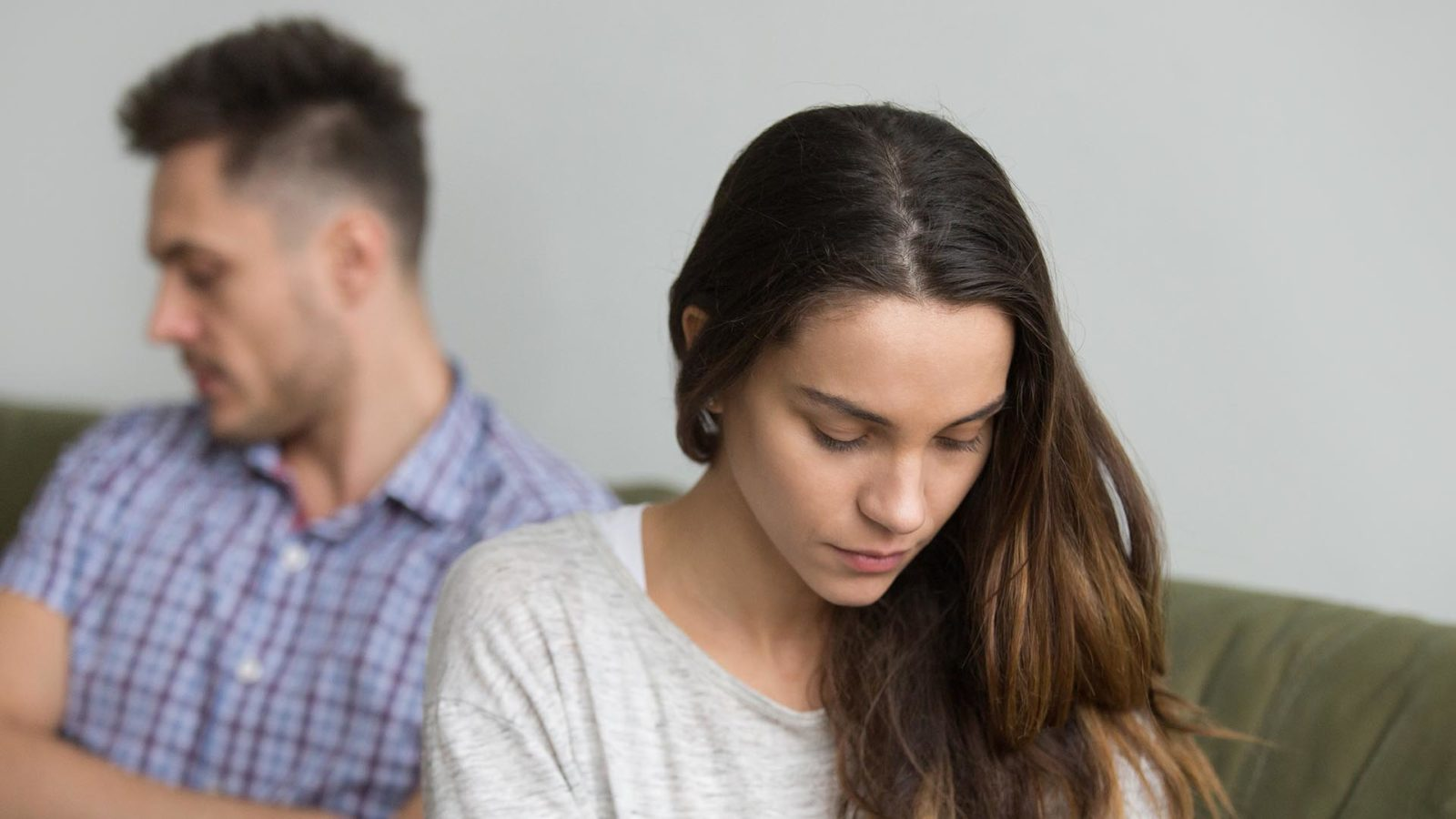 A young woman and man sitting back-to-back on a couch, both appearing upset and looking downward.