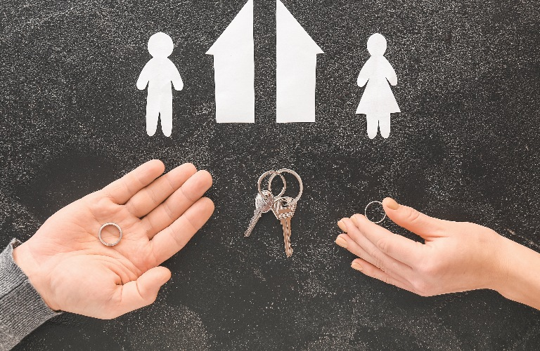 Hands holding wedding rings with paper cutouts of a man, woman, house, and keys on a black surface.