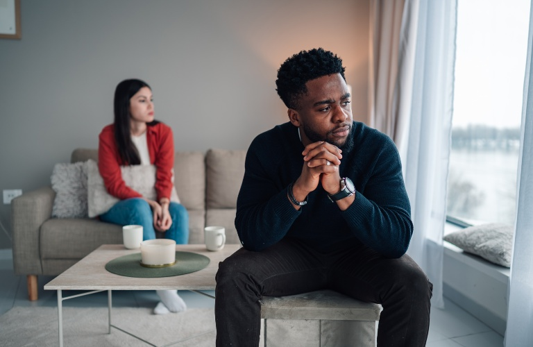 A man in a dark sweater sits pensively by a window while a woman in a red jacket sits on a couch behind him.