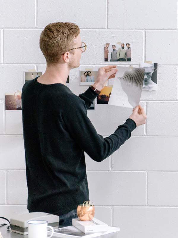 Man in black sweater arranging printed photos on white brick wall in bright room.