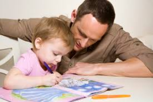 Adult male assisting young child with coloring in a book at a white table.