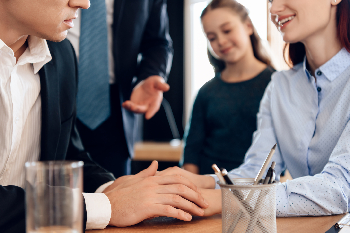 Two adults holding hands across a desk with a young girl and a businessman in the background.