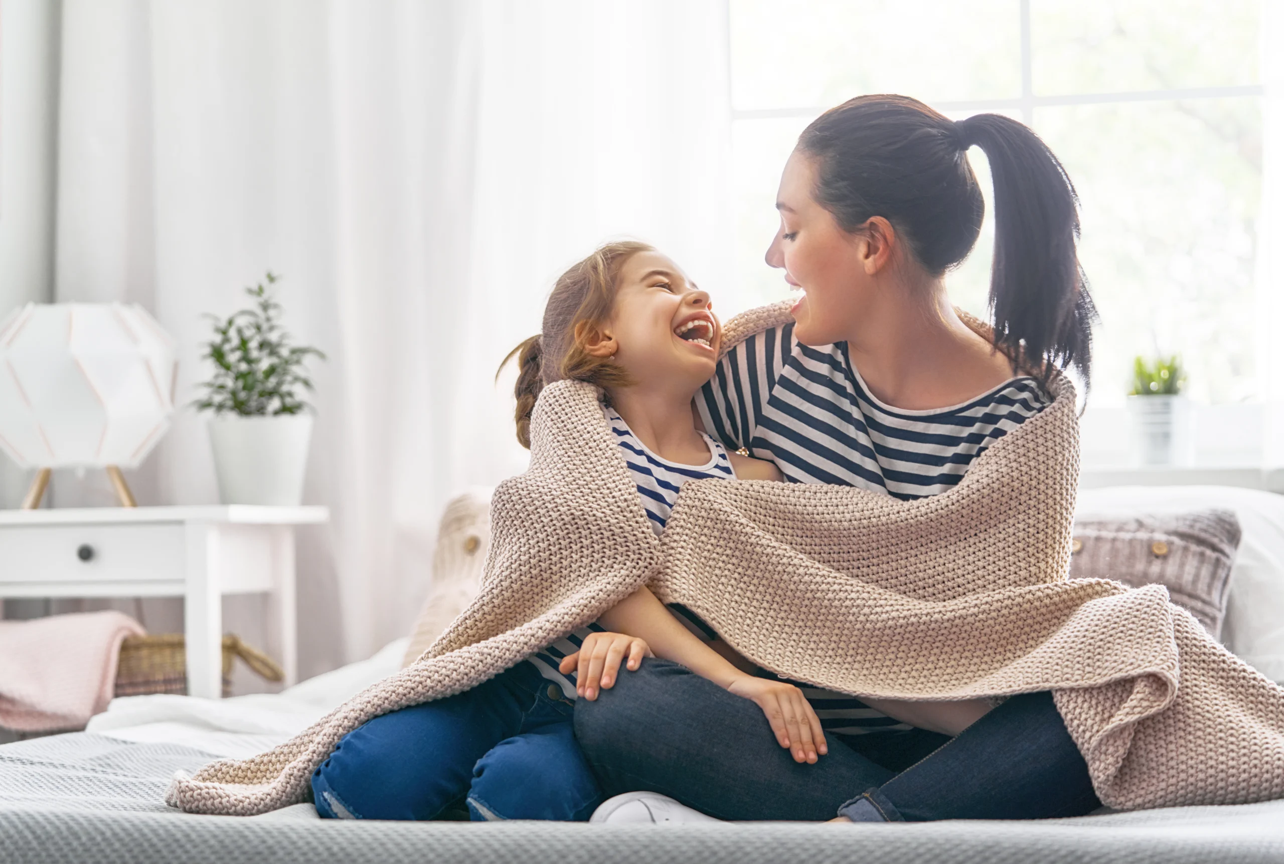 Mother and daughter wrapped in beige blanket, smiling and laughing indoors.
