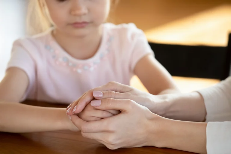 Close-up of an adult gently holding a child's hands across a wooden table.
