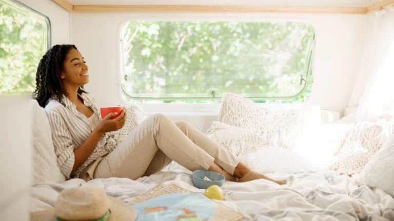 Woman in beige pants and striped shirt sitting on bed inside camper, holding red mug.