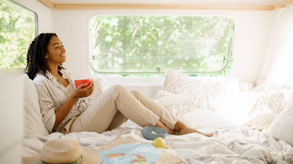Woman in beige pants and striped shirt sitting on bed inside camper, holding red mug.