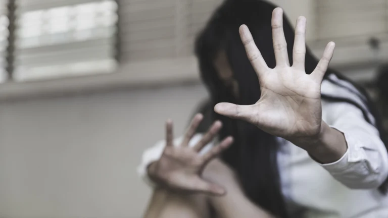 Person with long dark hair and white shirt raising hands defensively in muted indoor setting.