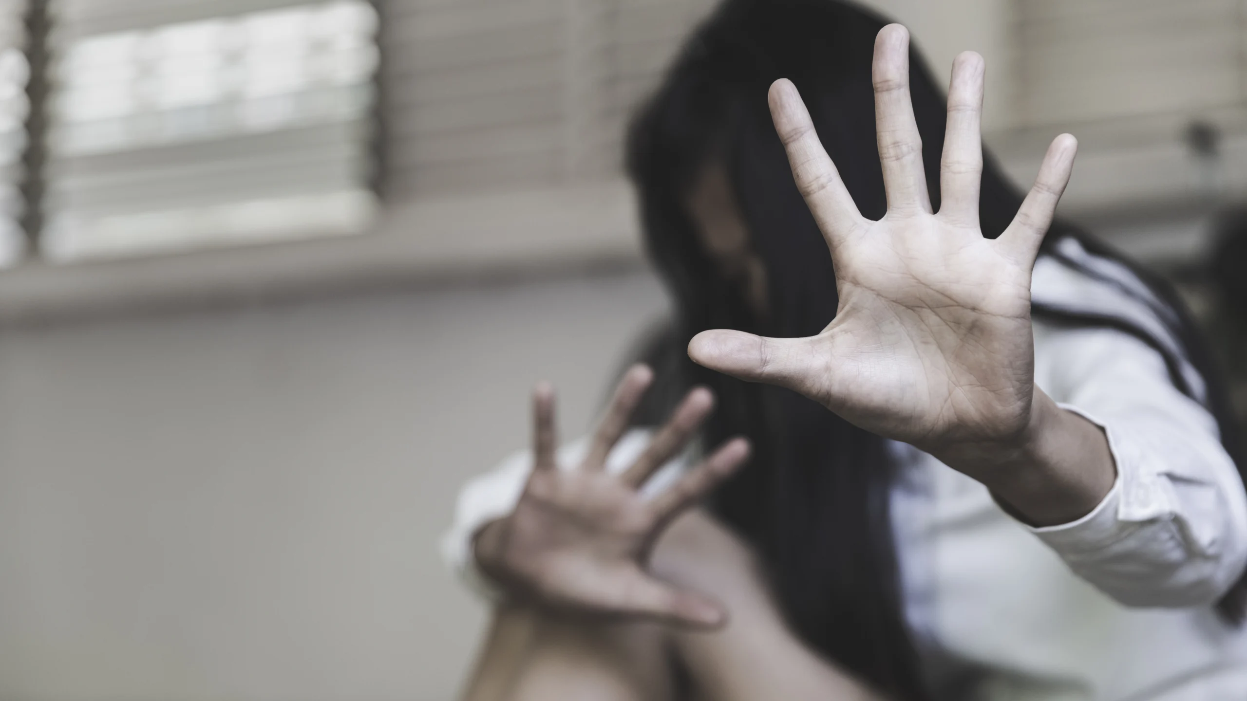 Person with long dark hair and white shirt raising hands defensively in muted indoor setting.