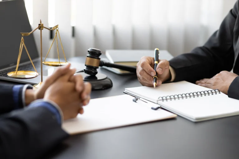 Two professionals in suits at a desk with legal scales, gavel, notebook, and pen.