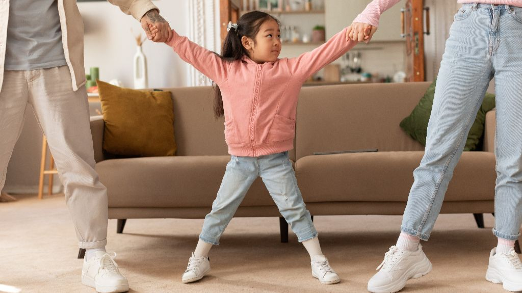 Young girl in pink jacket holding hands with two adults in casual attire indoors.