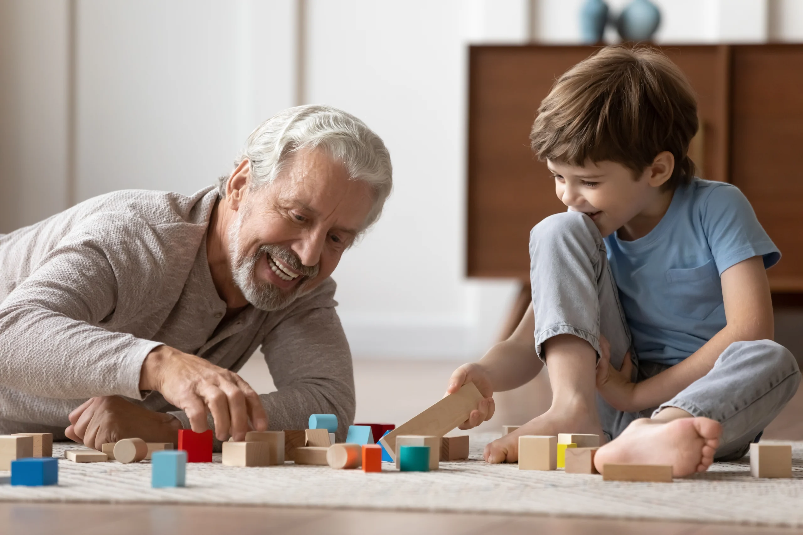 Elderly man and young boy playing with colorful wooden blocks on carpet indoors.