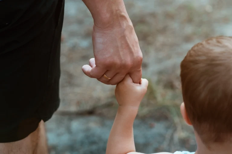Close-up of an adult holding a child's hand outdoors, adult wearing a wedding ring.