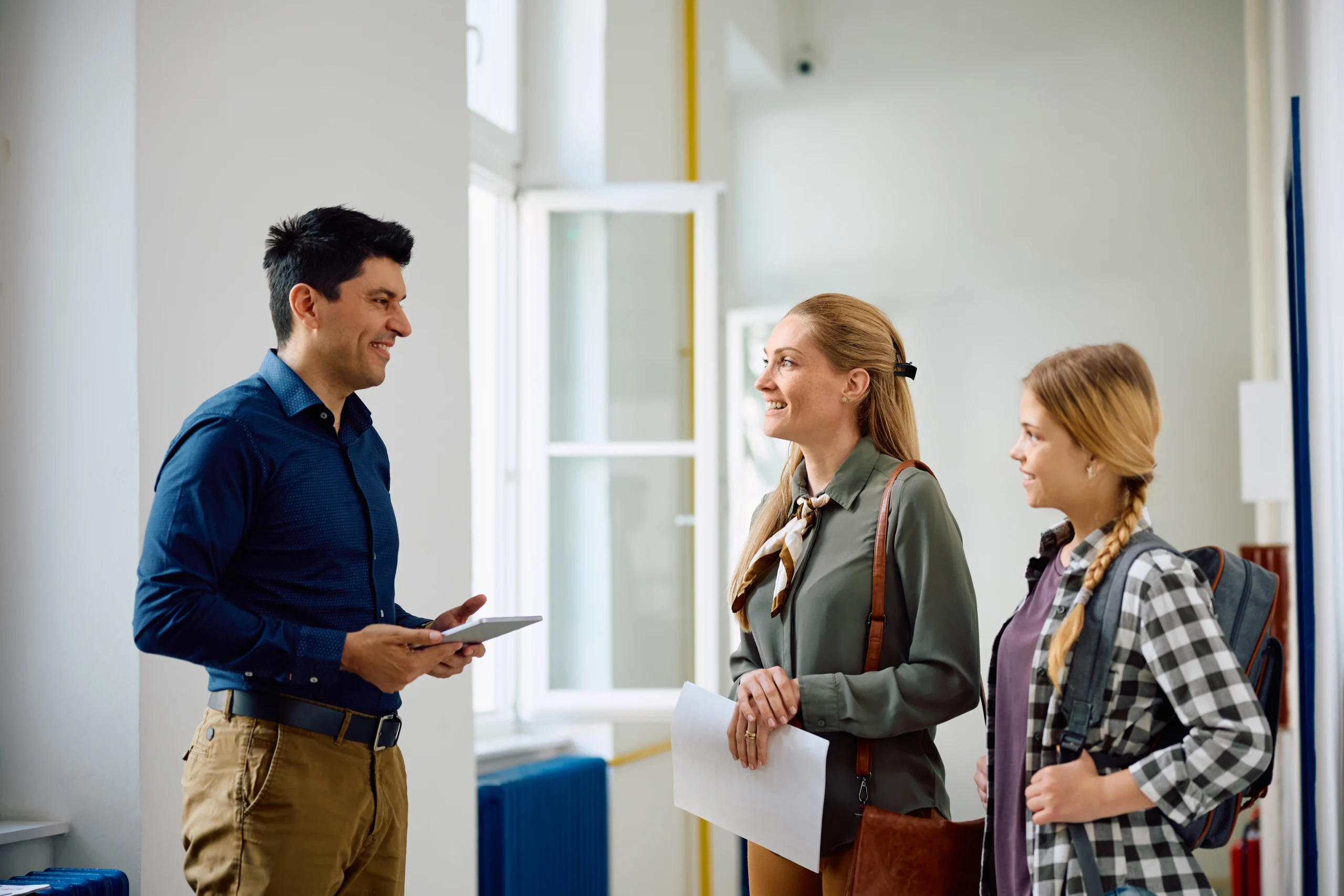 Three people engaged in conversation in a bright hallway, one man holding a tablet, two women smiling.