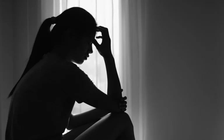 Silhouette of a woman sitting with hand on forehead, appearing distressed, near a window.