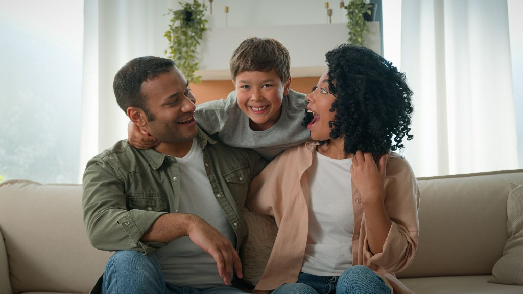 A smiling family of three sitting on a beige couch in a bright living room.