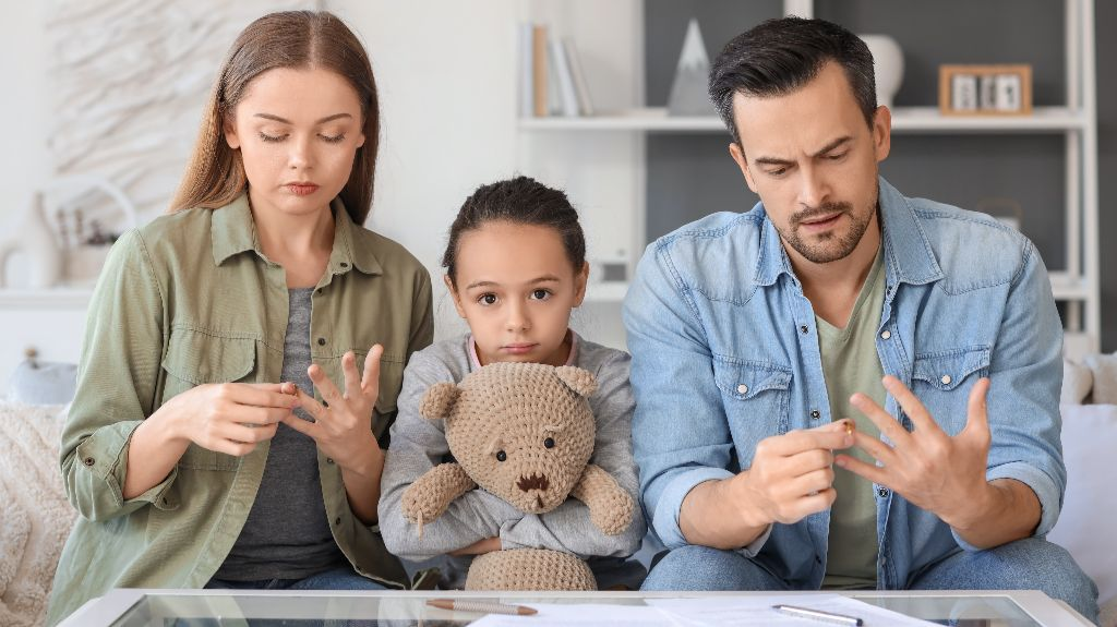 A family of three sitting on a couch, parents removing rings, child holding a teddy bear.
