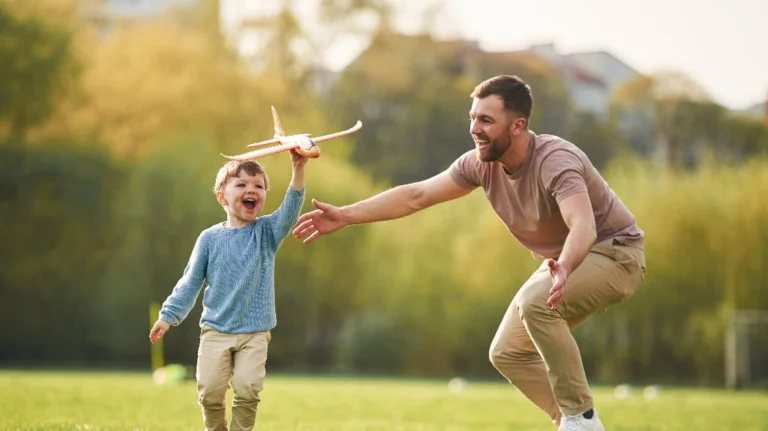Young boy holding a toy airplane with an adult male reaching out, outdoors in a green field.