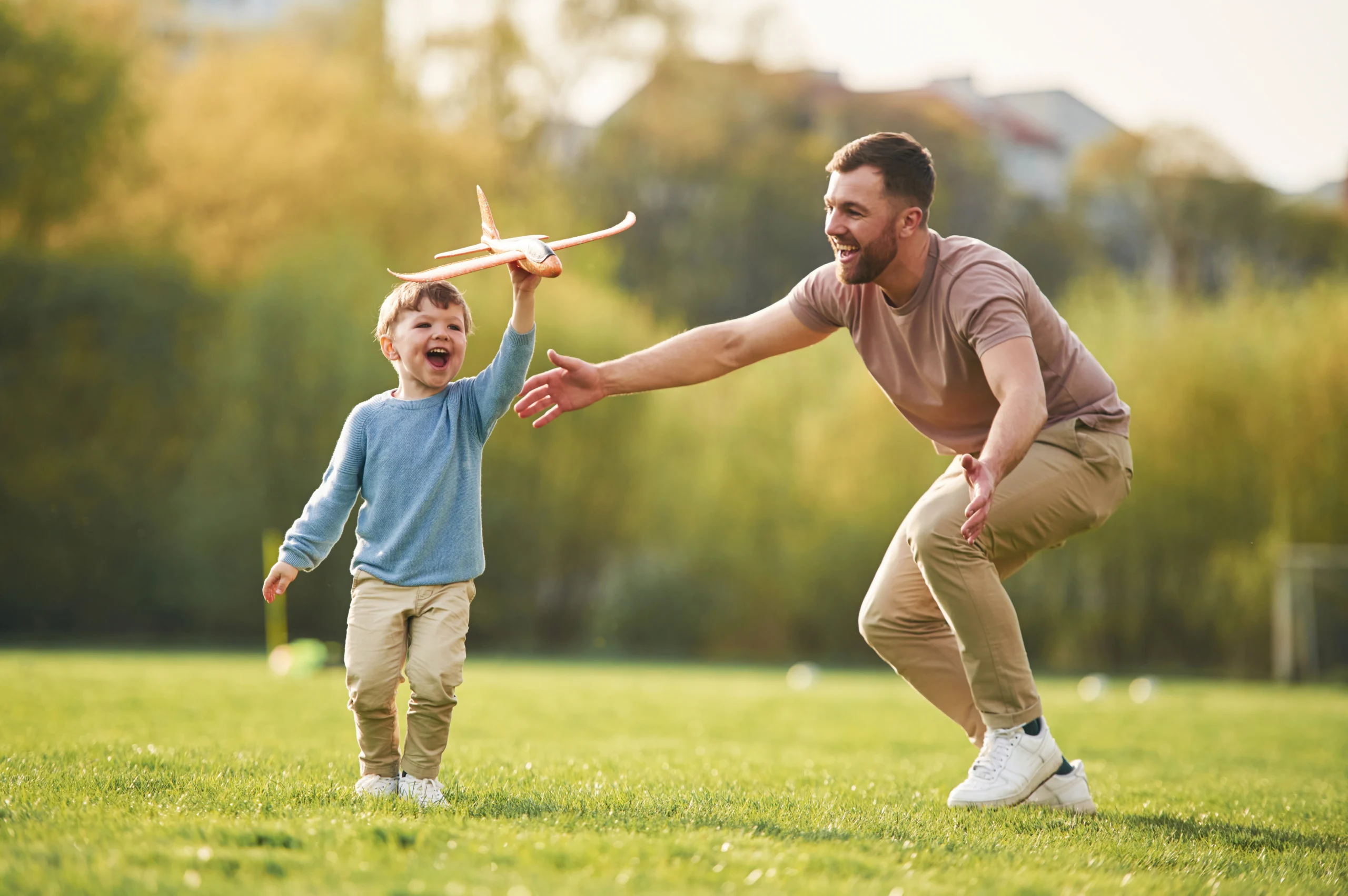 Young boy playing with toy airplane outdoors while man joyfully reaches out to him.