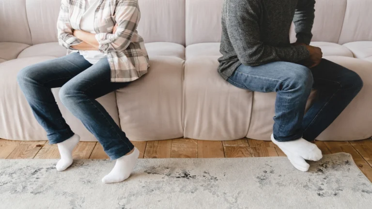 Two individuals sitting apart on a beige couch, facing away with crossed arms, wearing casual clothing and white socks.